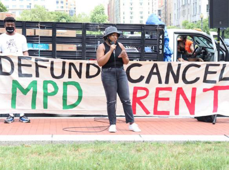 Photo of someone speaking into a microphone with banner saying 'Defund MPD, Cancel Rent'