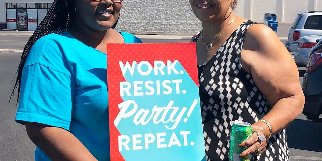 Two organizers holding a sign that says, "Work, Resist, Party, Repeat." Photo credit: Make It Work Nevada